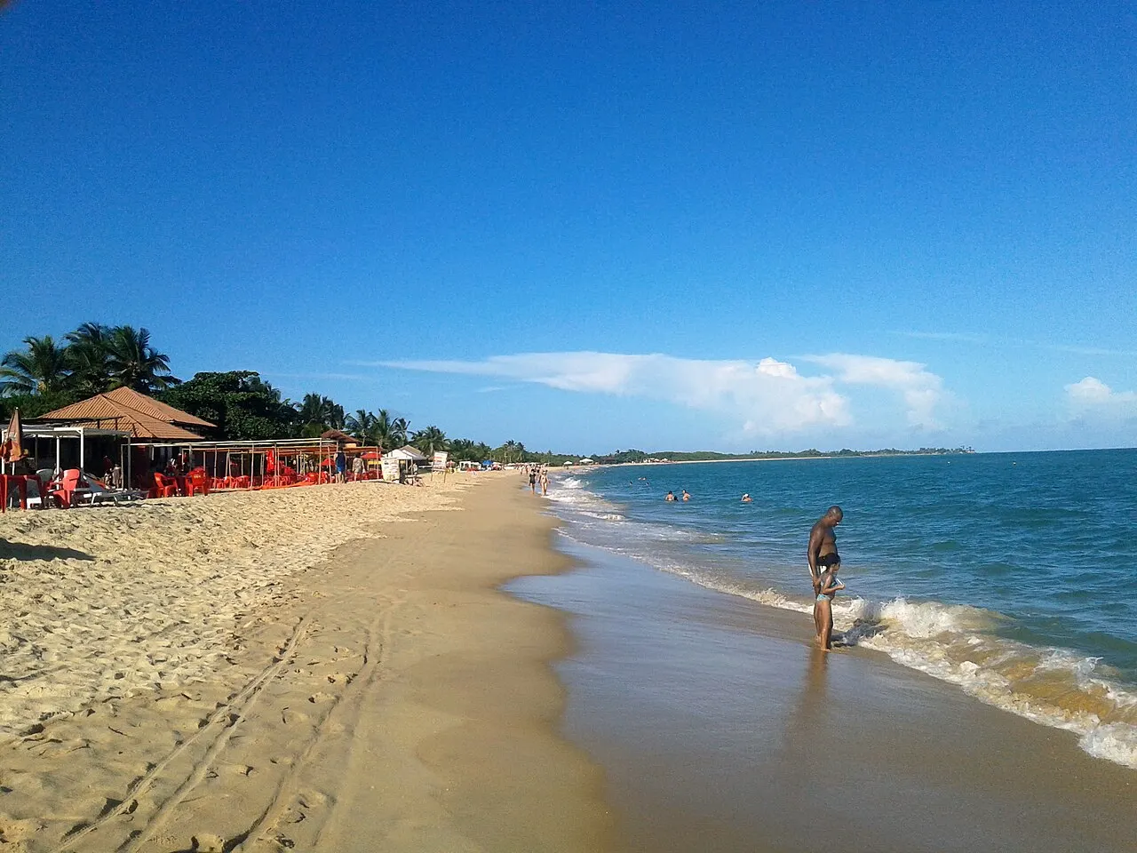A Praia de Taperapuã, uma faixa de areia clara com barracas de cobertura vermelha ao fundo e o mar verde e calmo.