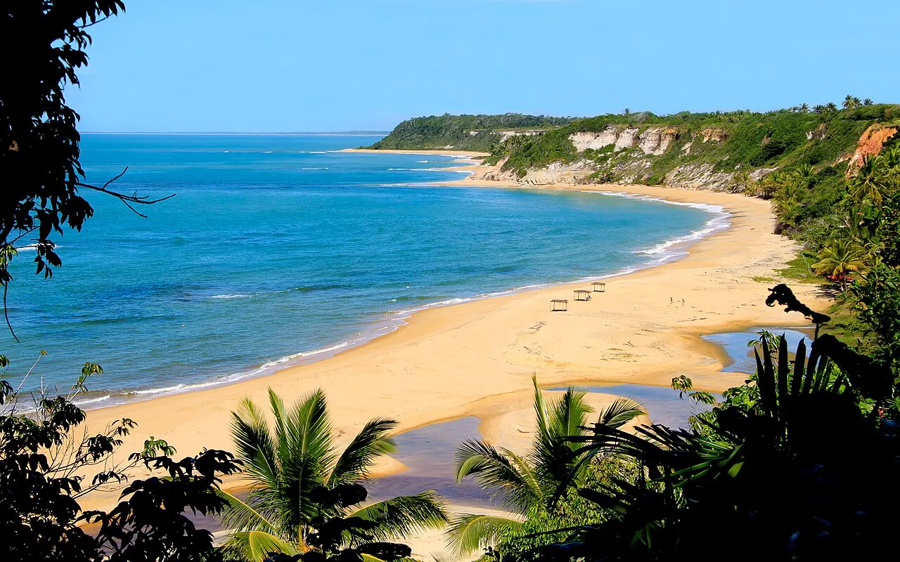 A Praia do Espelho vista do alto, uma curva de areia dourada entre a água azul-turquesa e falésias cobertas de mata.