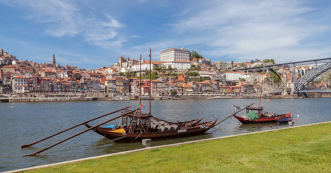 Barcos rabelos de madeira carregados com barris de vinho ancorados no rio Douro, com o centro histórico do Porto e a ponte Dom Luís ao fundo sob céu azul.