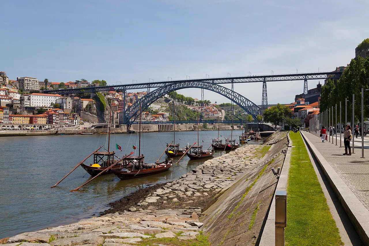 Ponte de ferro Luís I em arco sobre o rio Douro, com barcos rabelos ancorados na margem e casas coloridas subindo a encosta, sob céu azul claro.