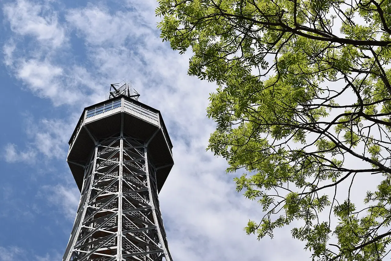 Petřín lookout tower seen from below, in open ironwork, with the glass top and tree leaves on the right.