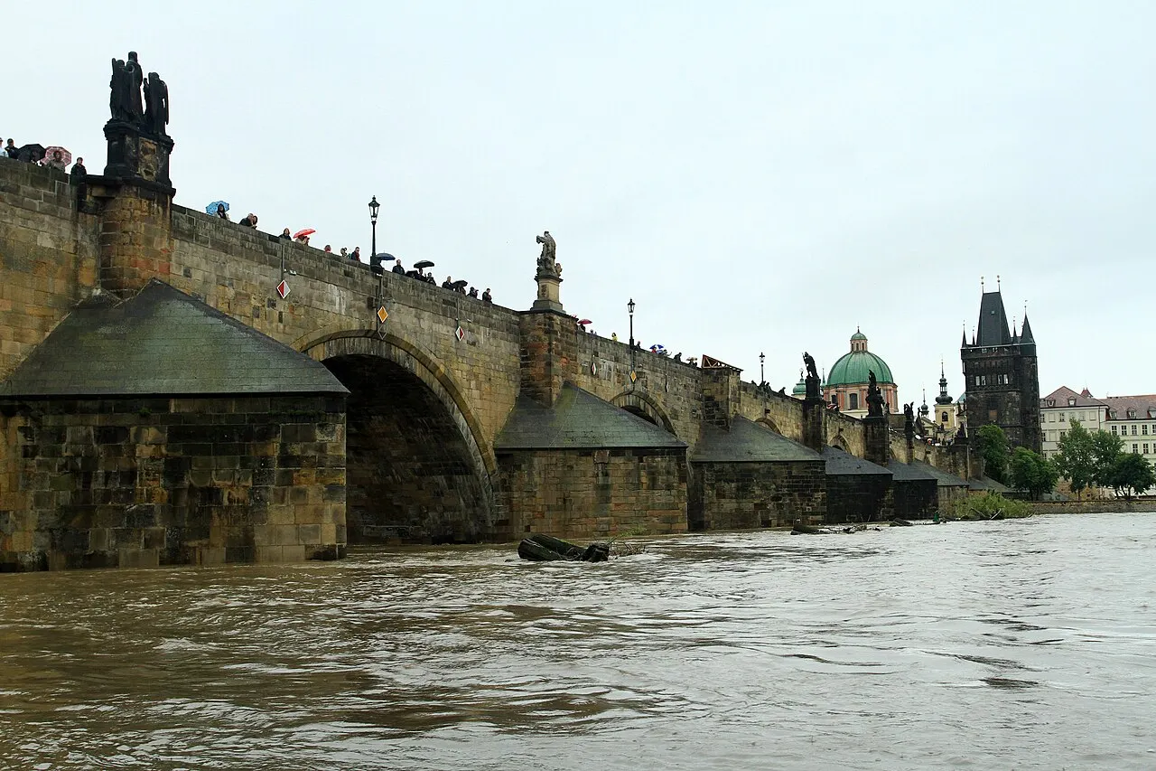 Charles Bridge with stone arches over the swollen, muddy Vltava River, statues on the parapet, people with umbrellas and Old Town towers in the background.