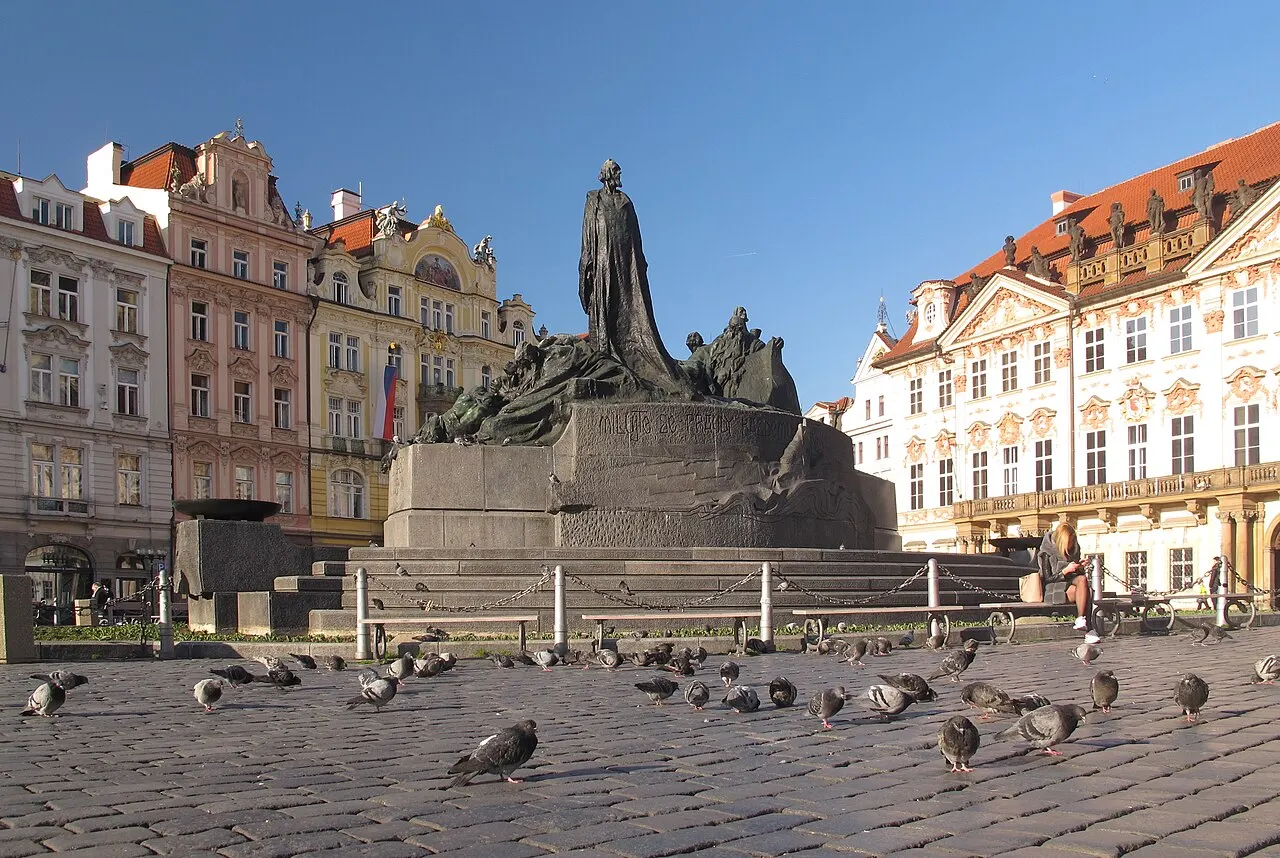 Monumento de bronze a Jan Hus, no meio da Praça da Cidade Velha de Praga, com as fachadas coloridas atrás.