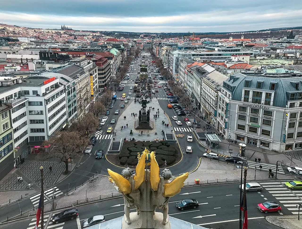 The view from above over Wenceslas Square running straight down between façades of every period, the equestrian statue at the centre and Prague Castle on the hill beyond.