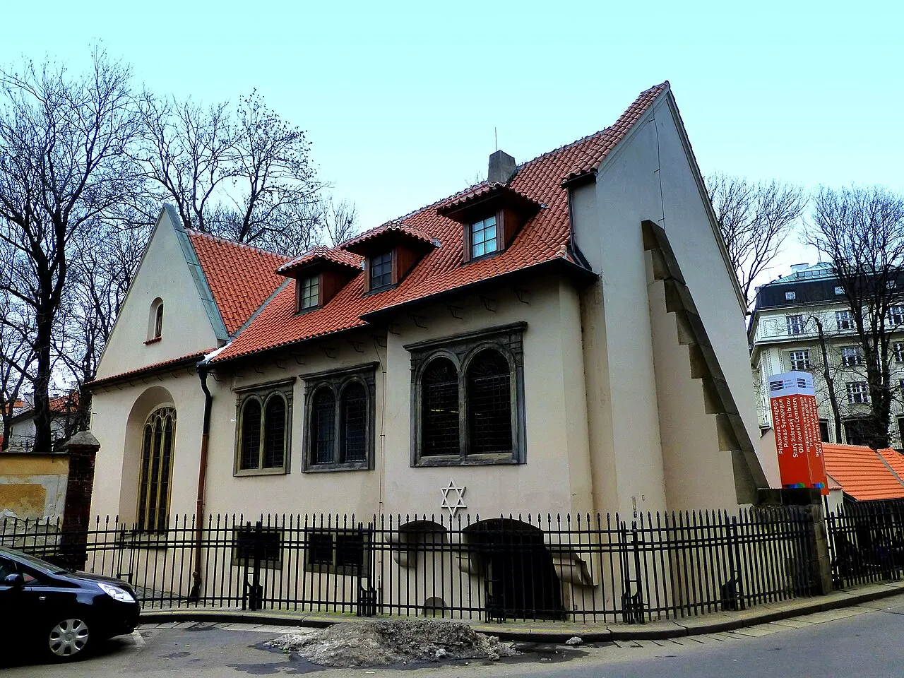Sinagoga Pinkas, em Praga, prédio claro de telhado vermelho, atrás de uma grade de ferro.