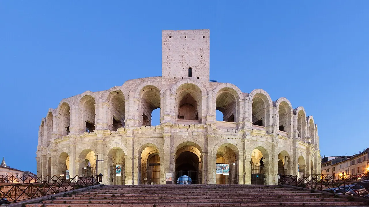 O anfiteatro romano de Arles com dois andares de arcos iluminados ao anoitecer, a torre medieval no alto e a escadaria na frente.