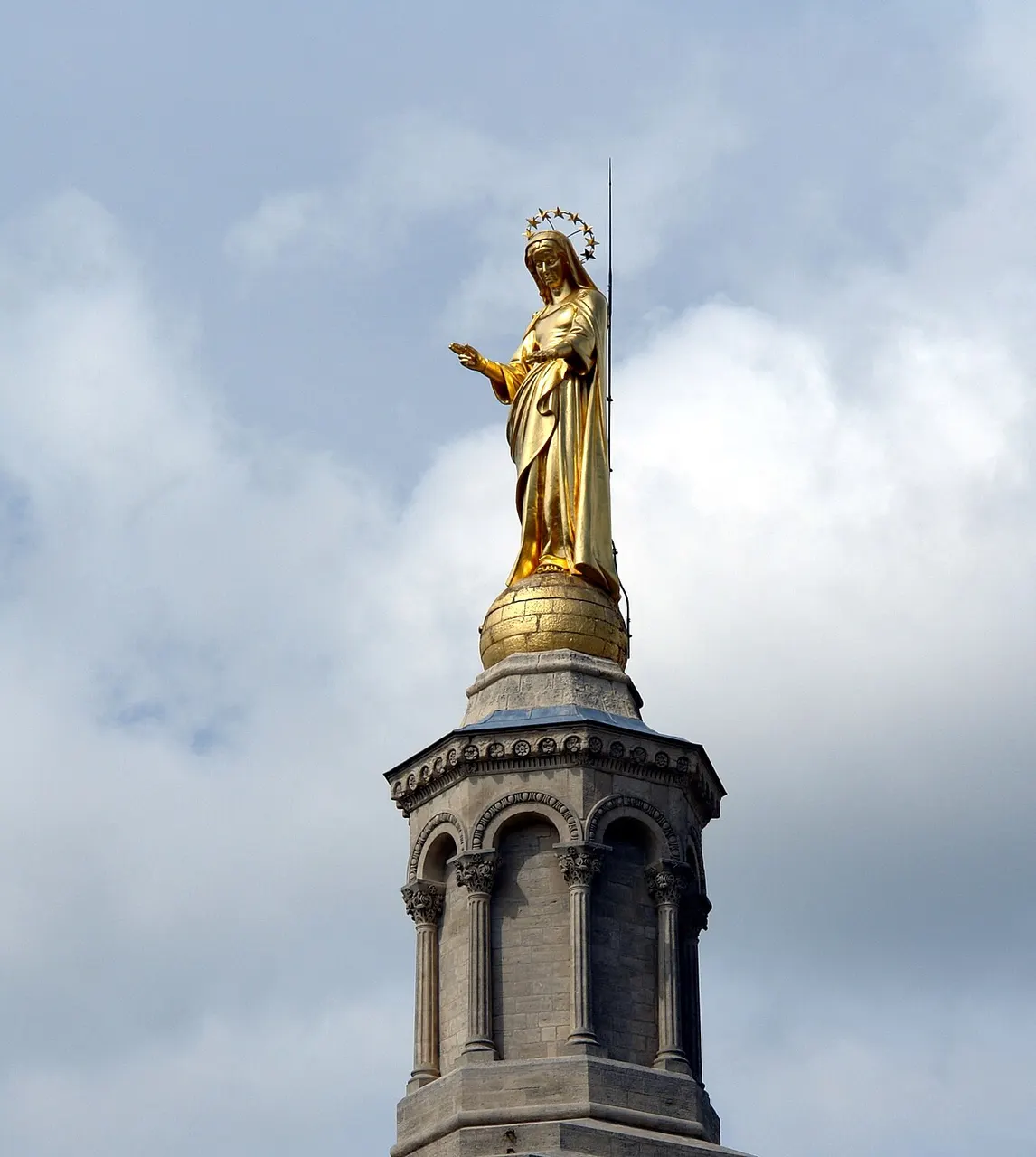 A estátua dourada da Virgem no alto do campanário de pedra da Catedral Notre-Dame des Doms, contra o céu nublado.