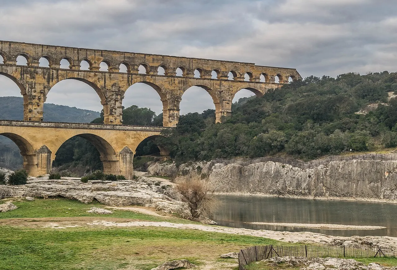 Os três níveis de arcos do aqueduto romano do Pont du Gard sobre o rio Gardon, com rochas claras e mata na margem.