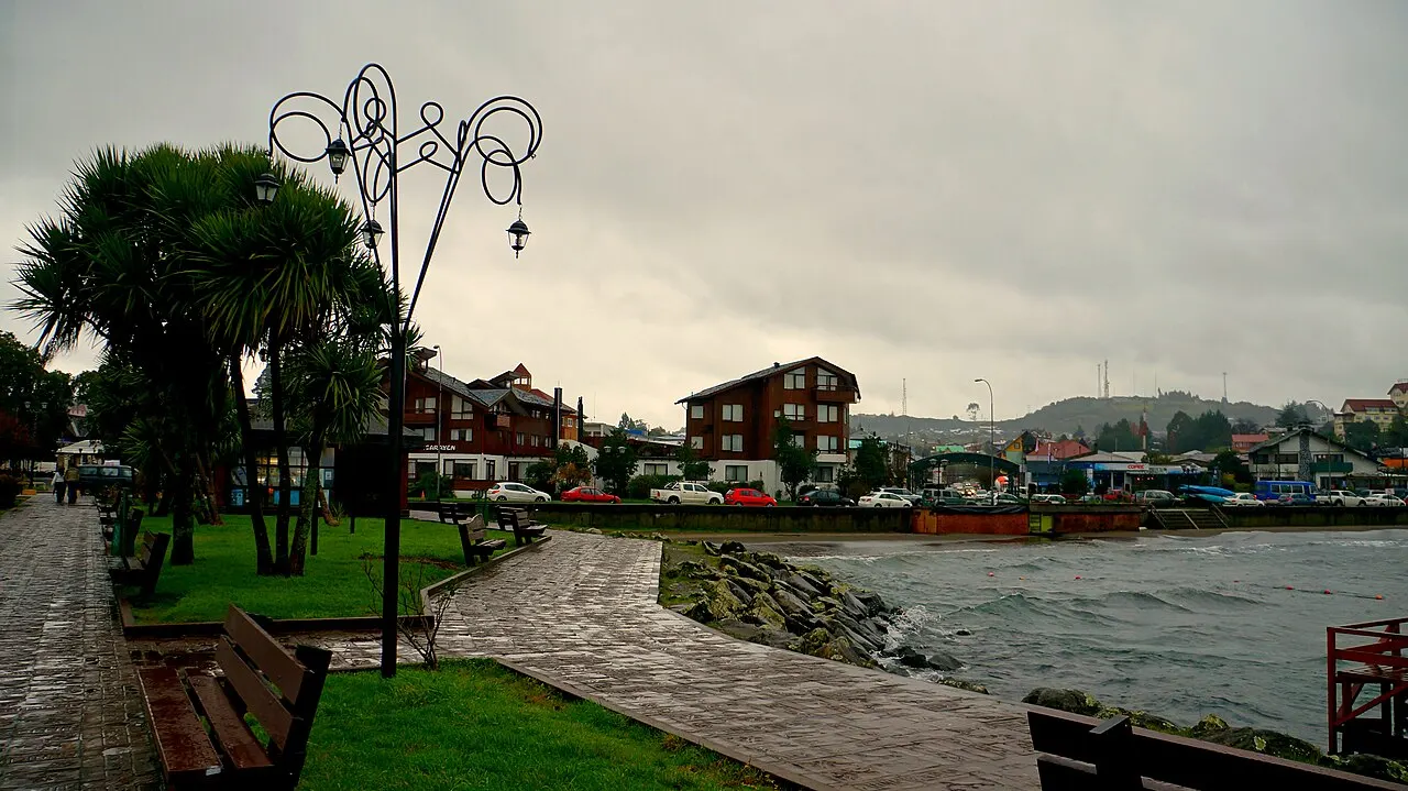 Calçadão de pedra molhado à beira do lago agitado, com bancos, palmeiras, um poste de luz ornamental e prédios de madeira ao fundo, sob céu nublado