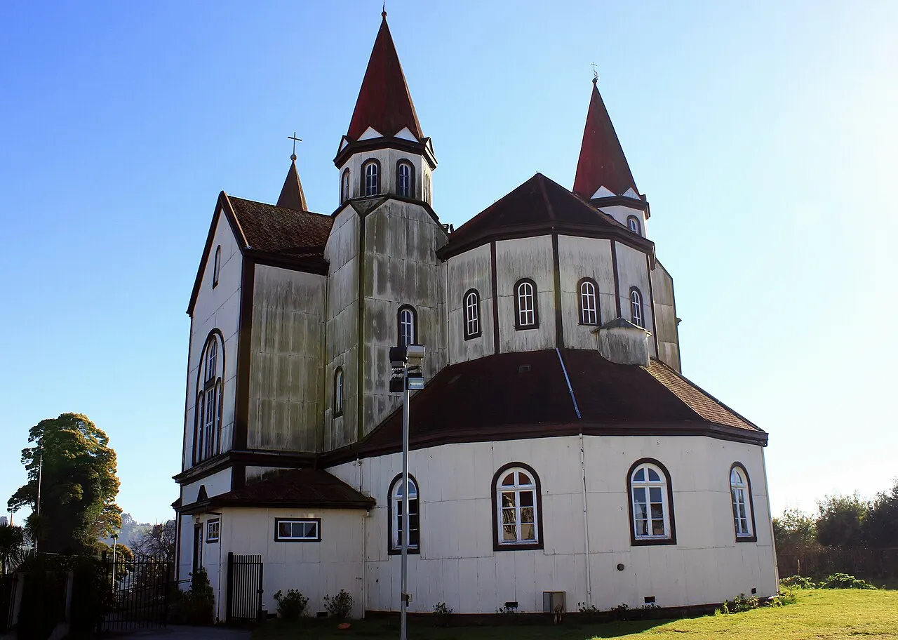 Igreja de madeira branca com telhados e torres pontiagudas vermelho-escuras, vista de trás sob céu azul, com gramado em primeiro plano