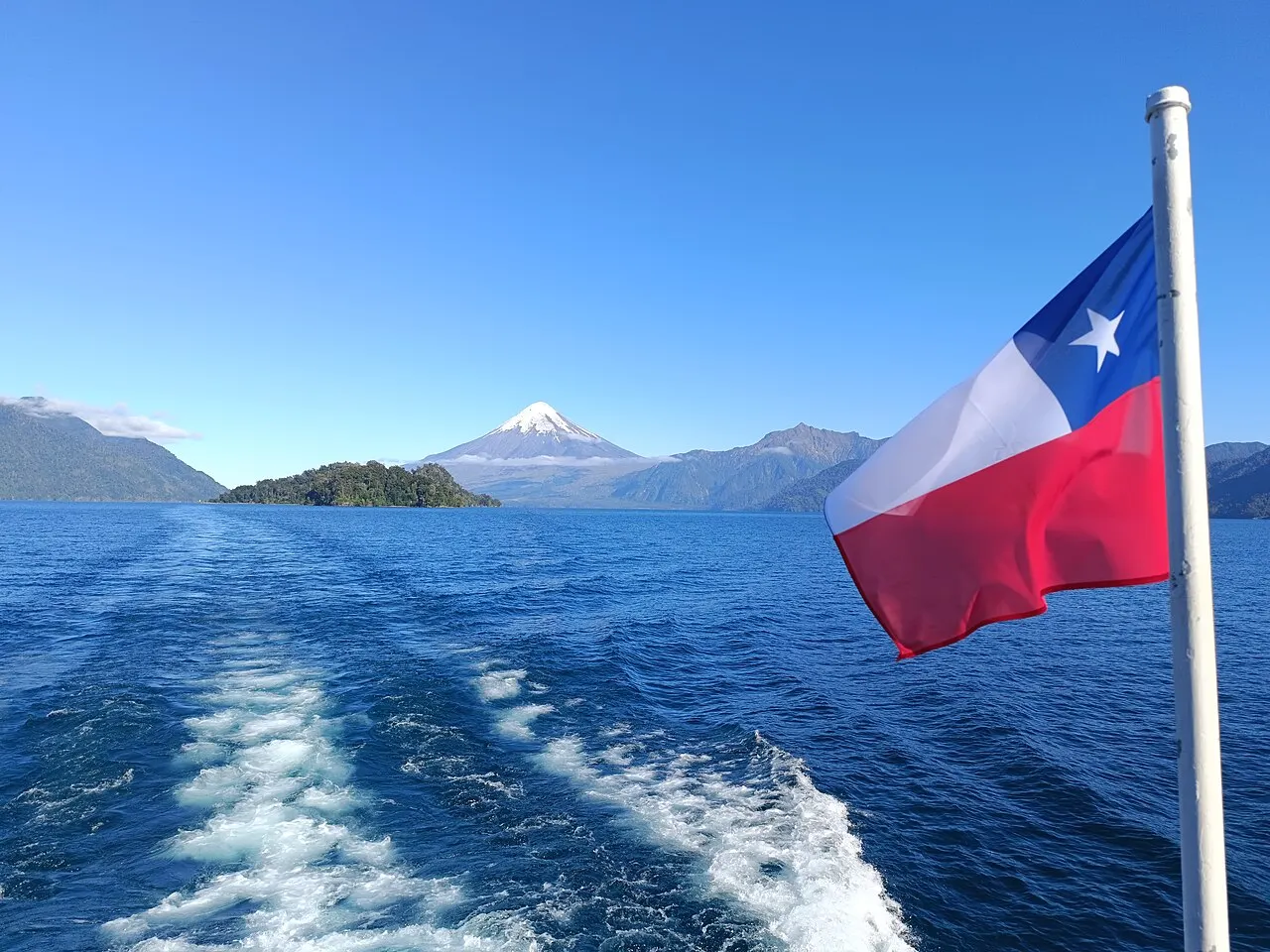 Esteira de um barco no lago azul, com a bandeira do Chile à direita, uma ilhota arborizada e um vulcão de cume nevado ao fundo