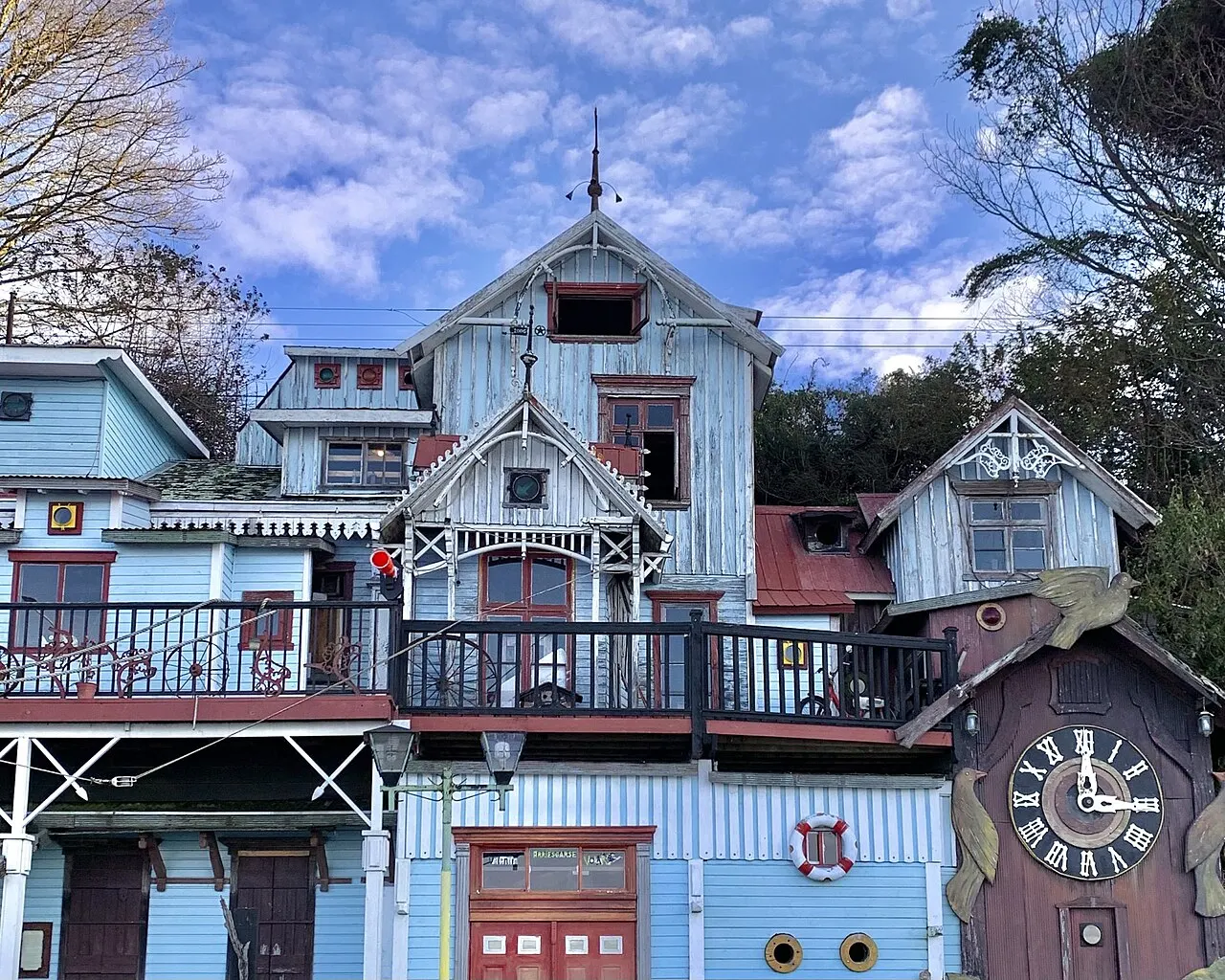 Fachada de madeira azul-clara com varanda de grade escura, boia salva-vidas, escotilhas e um grande relógio de cuco à direita