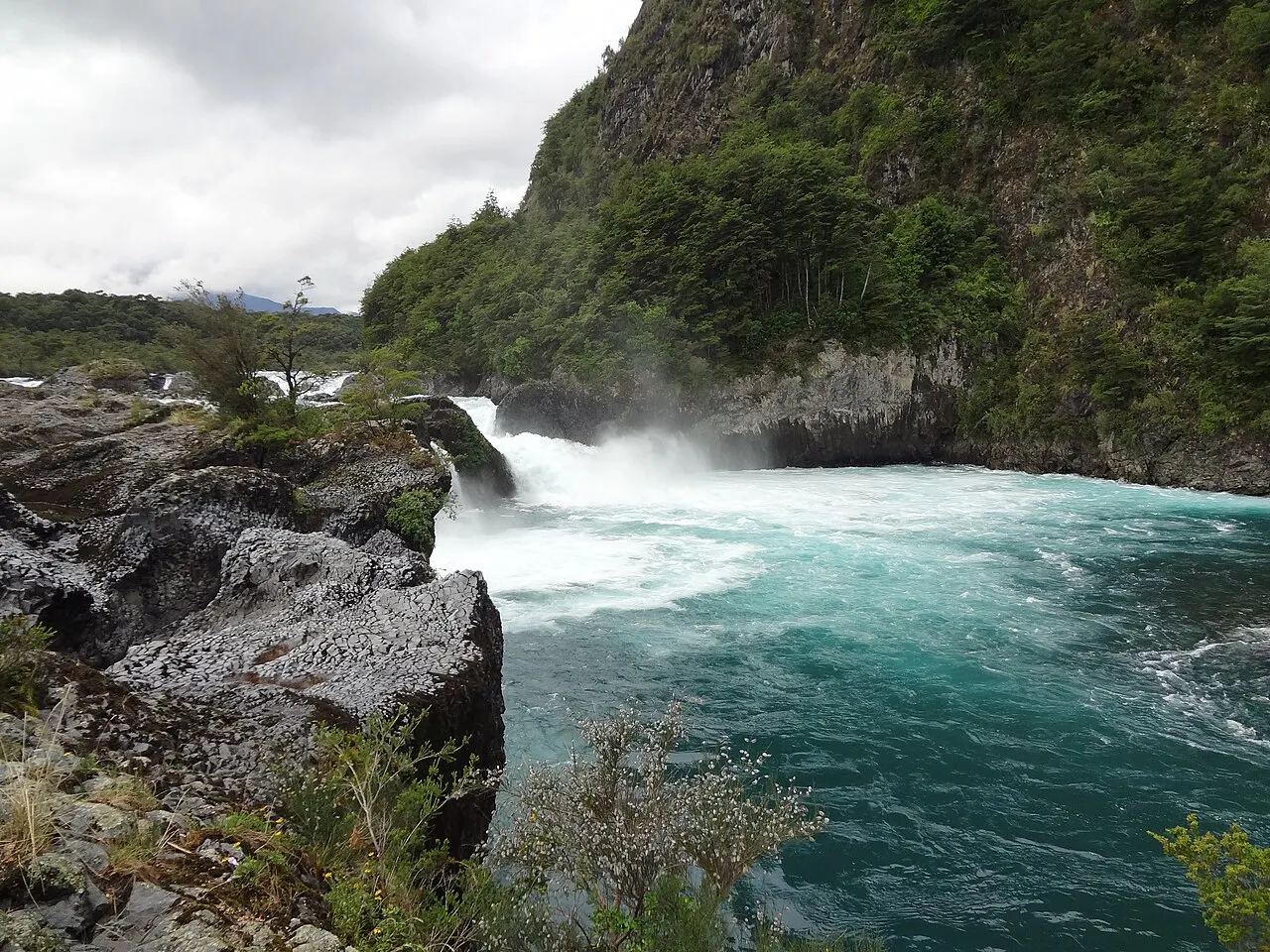 Água verde-azulada em espuma branca descendo entre rochas escuras de lava, ao pé de uma encosta coberta de mata, sob céu nublado