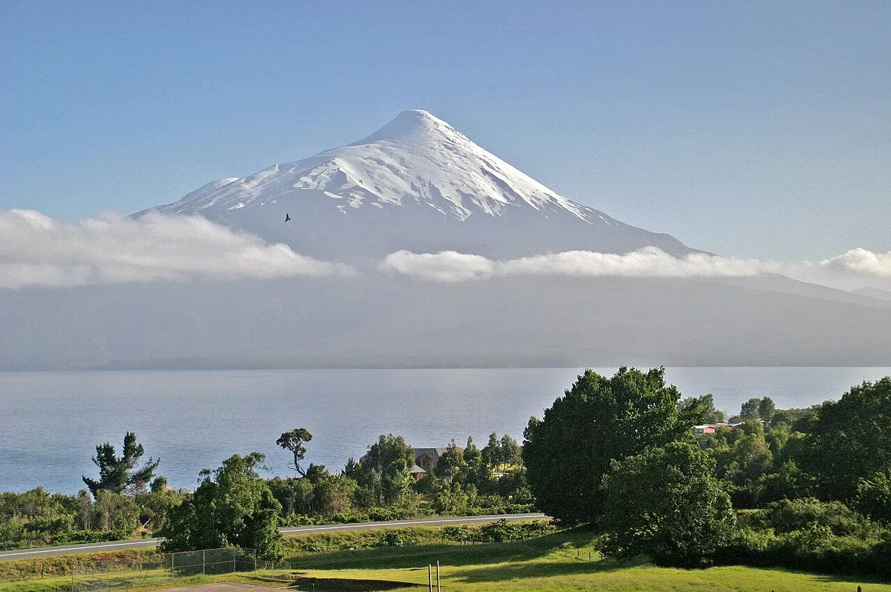 Vulcão em cone perfeito de cume nevado acima de uma faixa de nuvens, com um lago e árvores verdes em primeiro plano