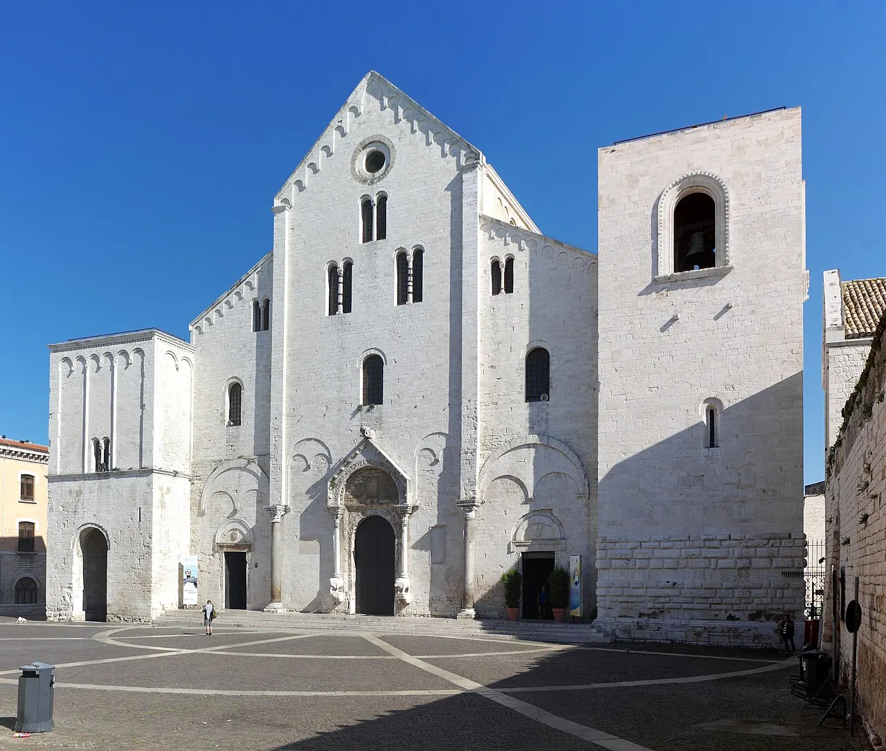 A fachada românica branca da Basílica de San Nicola, com a torre quadrada ao lado e a praça de pedra diante dela.