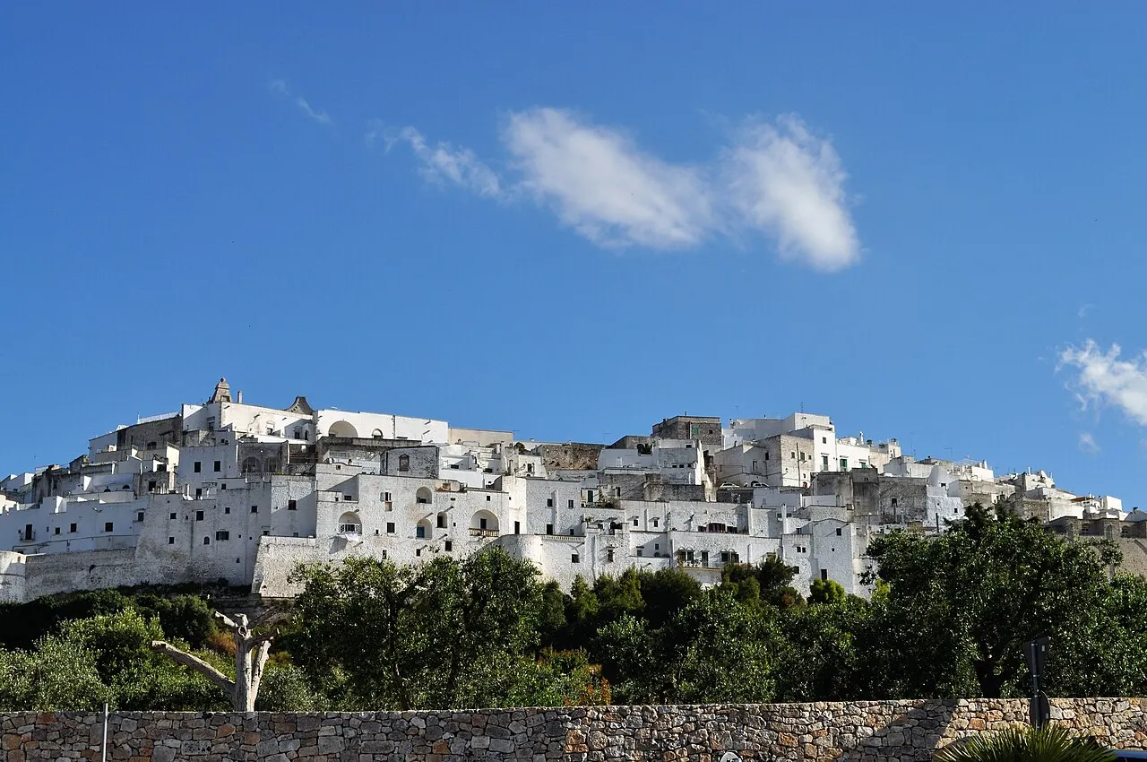 O casario branco de Ostuni cobrindo o alto da colina sob céu azul, com árvores e um muro de pedra embaixo.