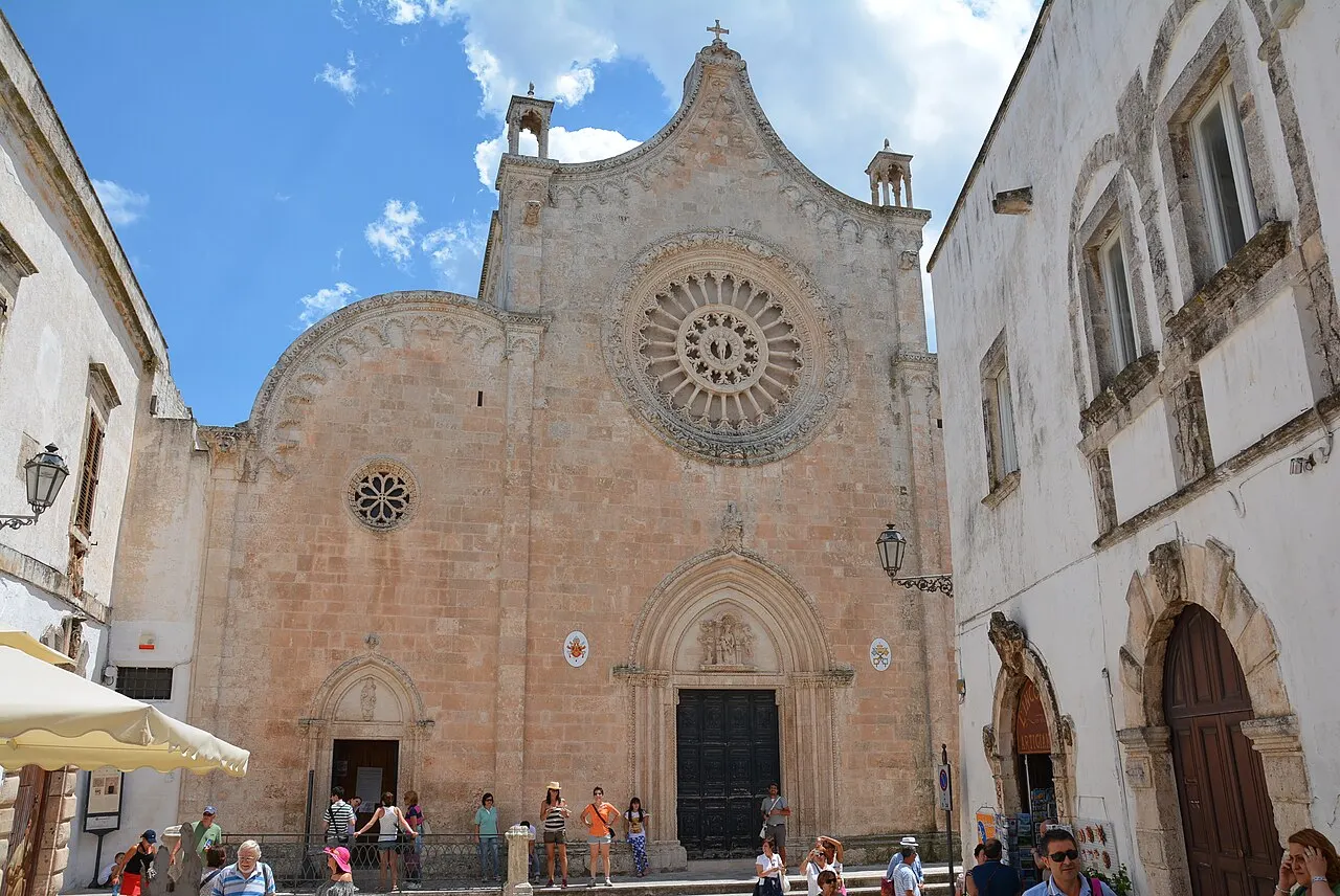 A fachada gótica da Catedral de Ostuni, com a grande rosácea, o frontão curvo e pessoas na praça diante dela.