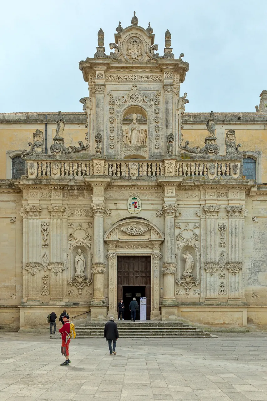 A fachada barroca do Duomo de Lecce, com a escadaria, a porta aberta e pessoas na Piazza del Duomo.