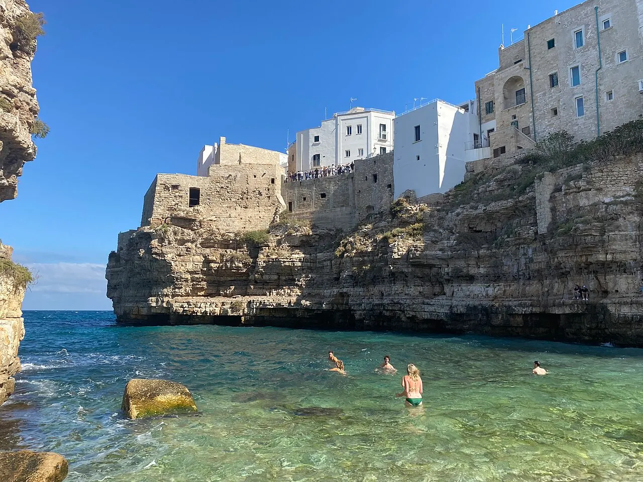 A água verde-clara da enseada de Lama Monachile com banhistas, entre falésias e as casas brancas de Polignano no alto.
