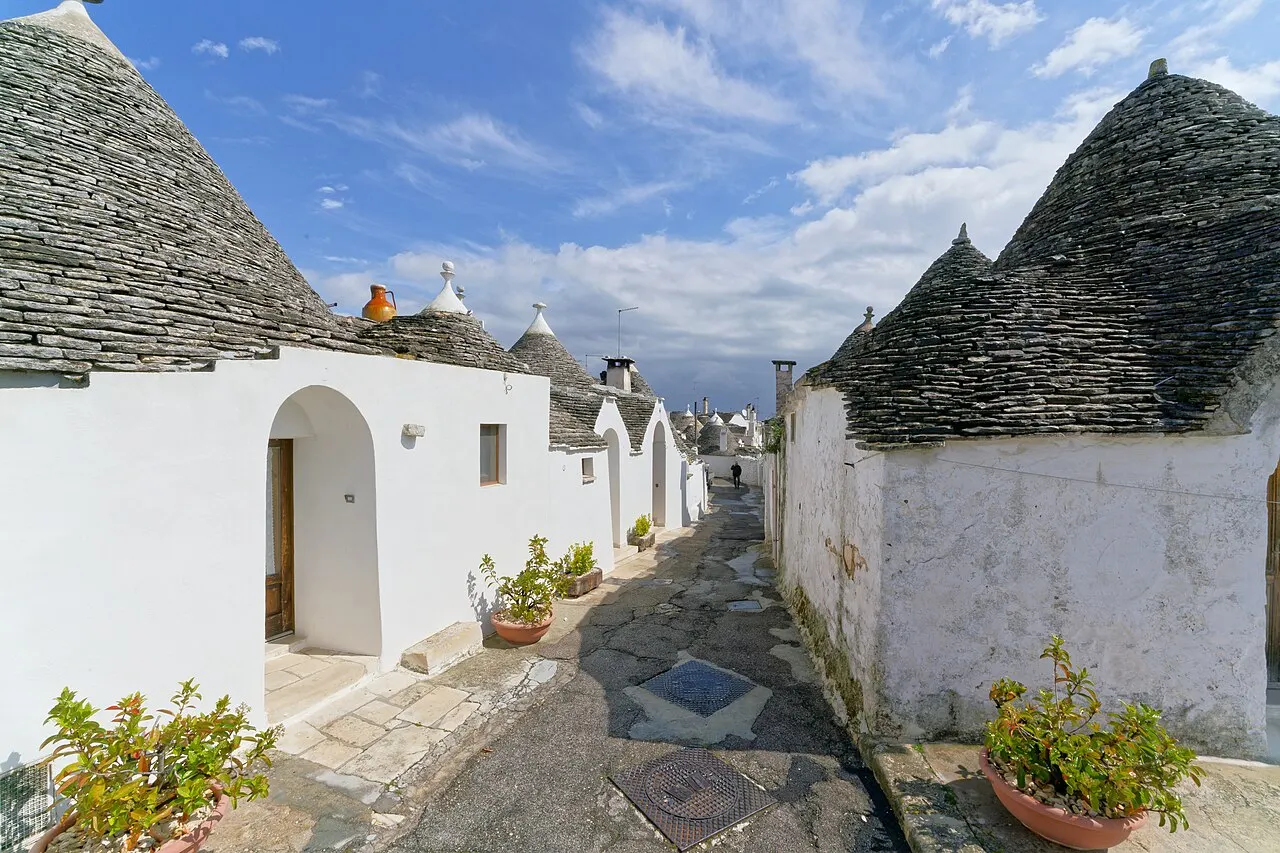 Uma ruela de pedra entre trulli brancos com telhados cônicos de pedra cinza e vasos de flores, no Rione Monti.