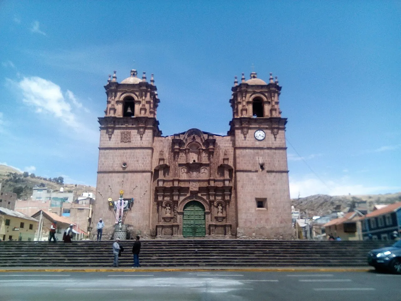 A fachada de pedra da catedral, com duas torres de sino, portada esculpida e porta verde, no alto de uma escadaria larga.