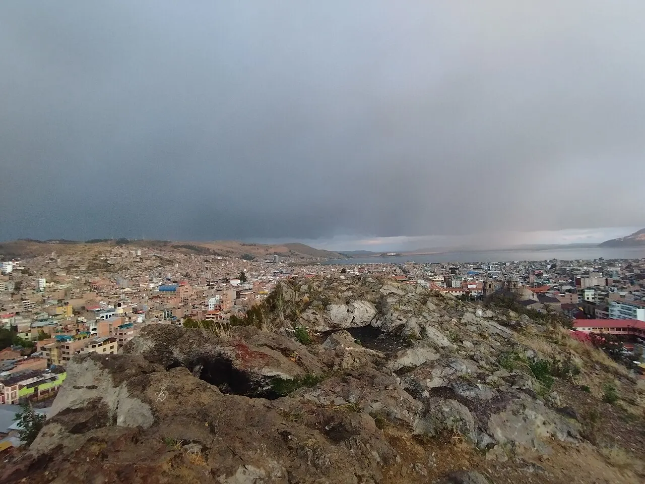 Rochas do alto do morro em primeiro plano e, abaixo, a cidade de Puno se estendendo até o lago sob céu nublado.