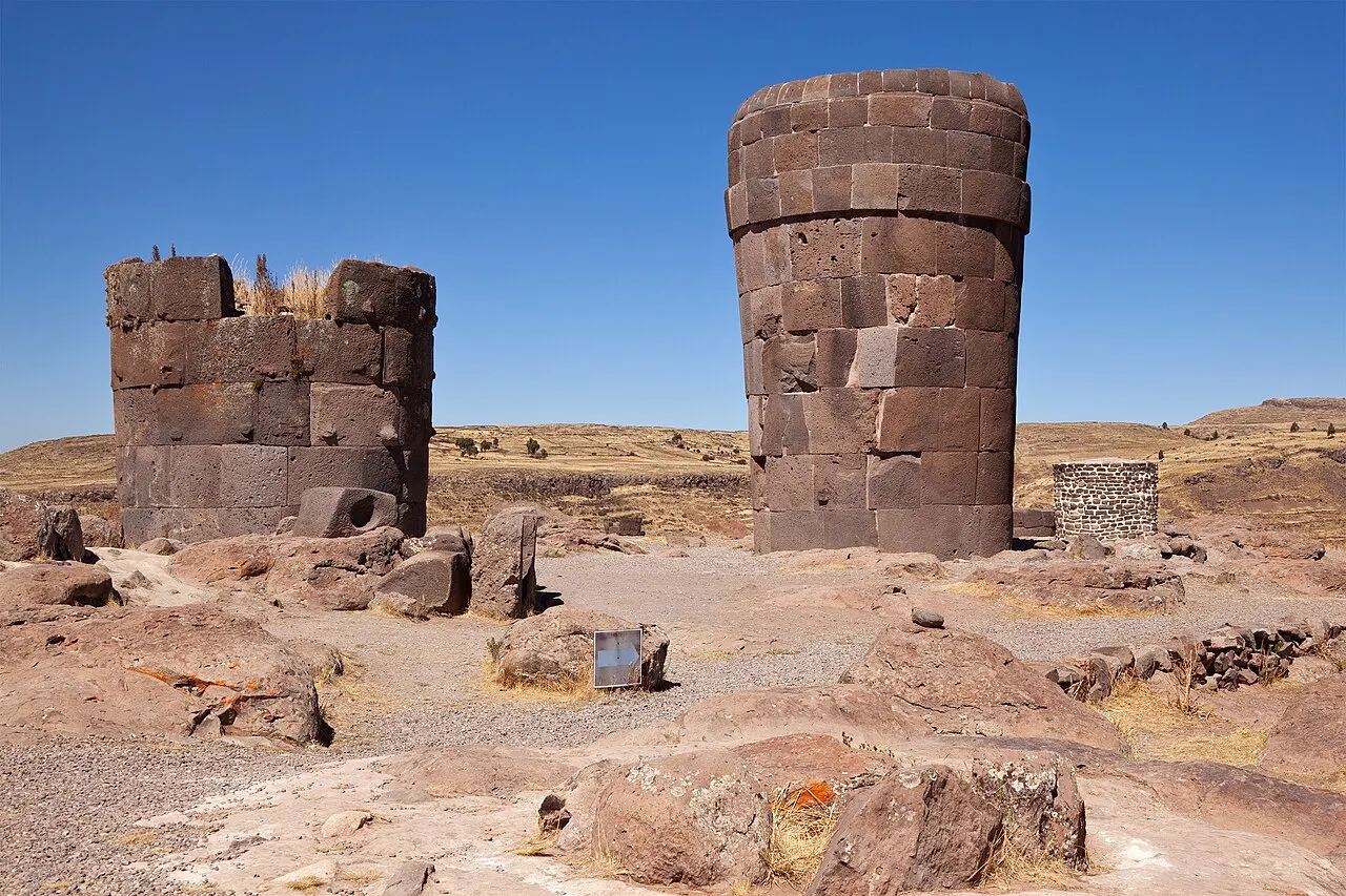Duas torres funerárias cilíndricas de blocos de pedra, uma inteira e uma mais baixa e partida, sobre chão de rocha avermelhada.