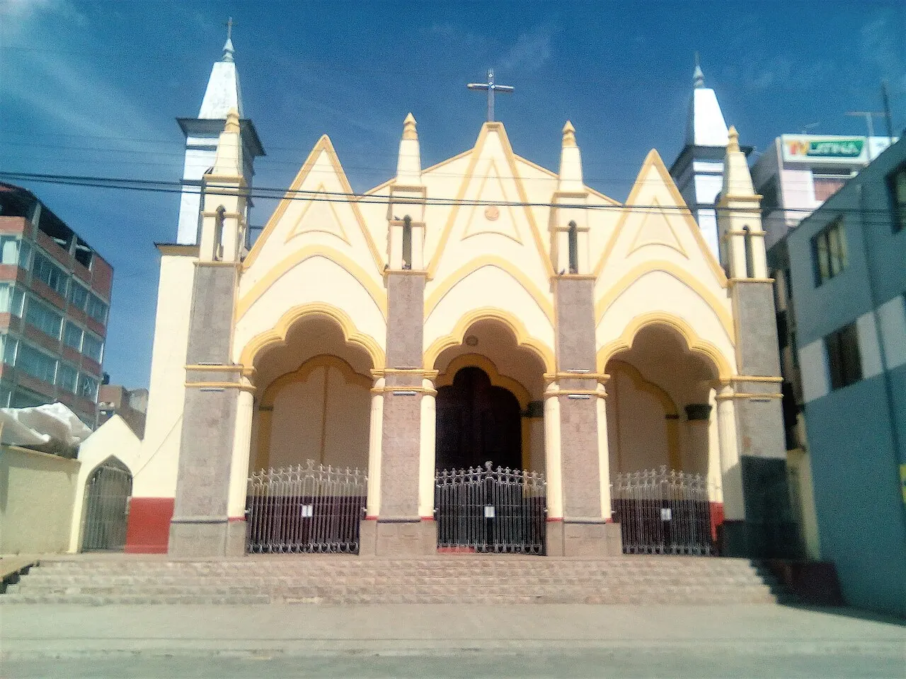 Uma igreja de fachada creme e amarela com três arcos de entrada gradeados, frontões pontudos e duas torres finas brancas.