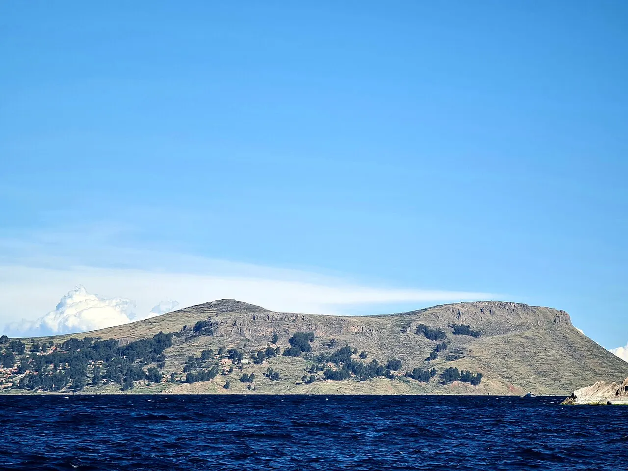 A ilha de Amantaní, com encostas em terraços e árvores na base, vista do lago azul-escuro sob céu limpo.