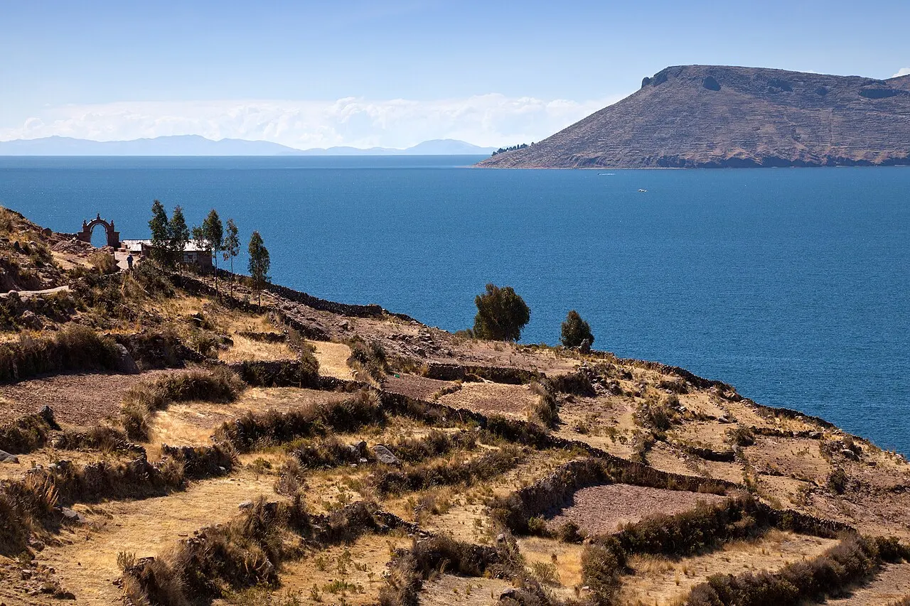 Terraços cercados por muros de pedra descem até o lago azul; no alto há um arco de pedra e árvores, e outra ilha ao fundo.
