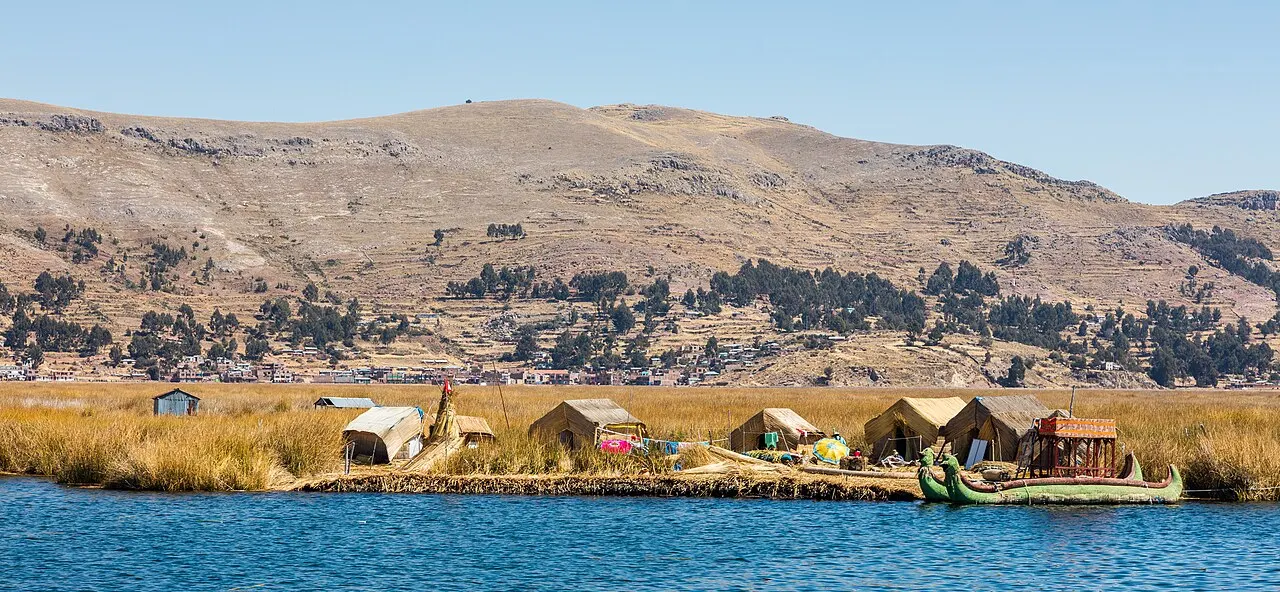 Uma ilha baixa de totora com casas de junco e barcos de proa alta na beira da água azul, e uma colina seca ao fundo.