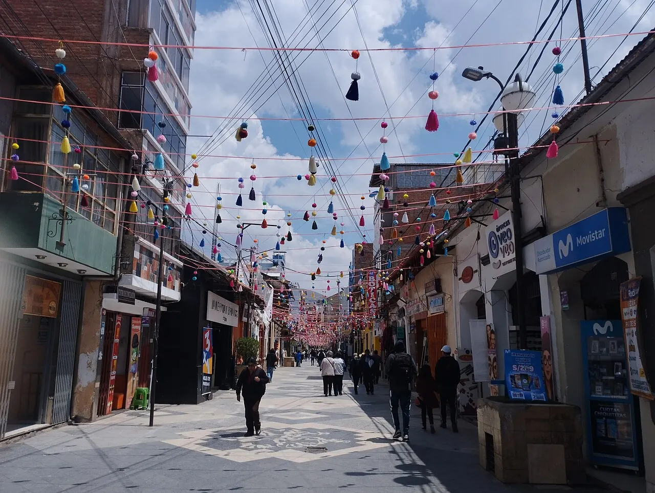 Uma rua de pedestres com lojas dos dois lados e fios cheios de pompons coloridos pendurados de um prédio ao outro.