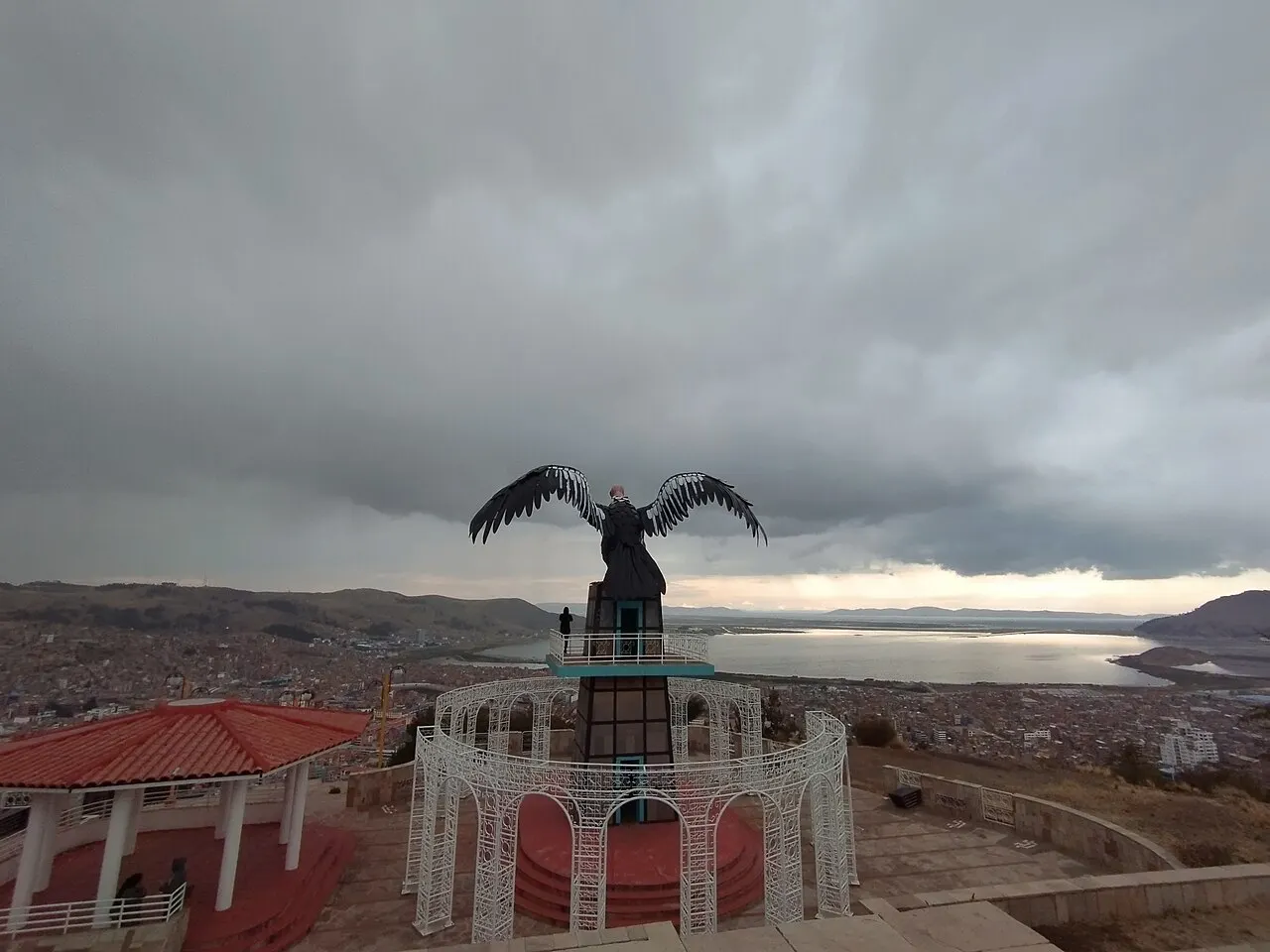 Uma estátua escura de condor de asas abertas sobre um pedestal no mirante, com a cidade e a baía do lago ao fundo sob nuvens.