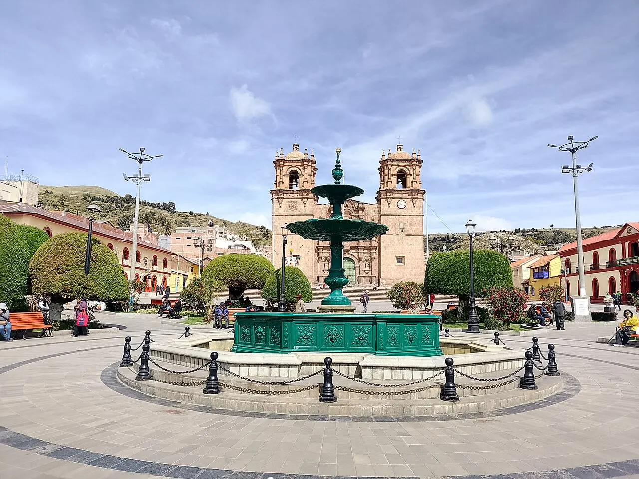 Um chafariz verde cercado por correntes no meio da praça, árvores podadas em volta e as duas torres da catedral ao fundo.