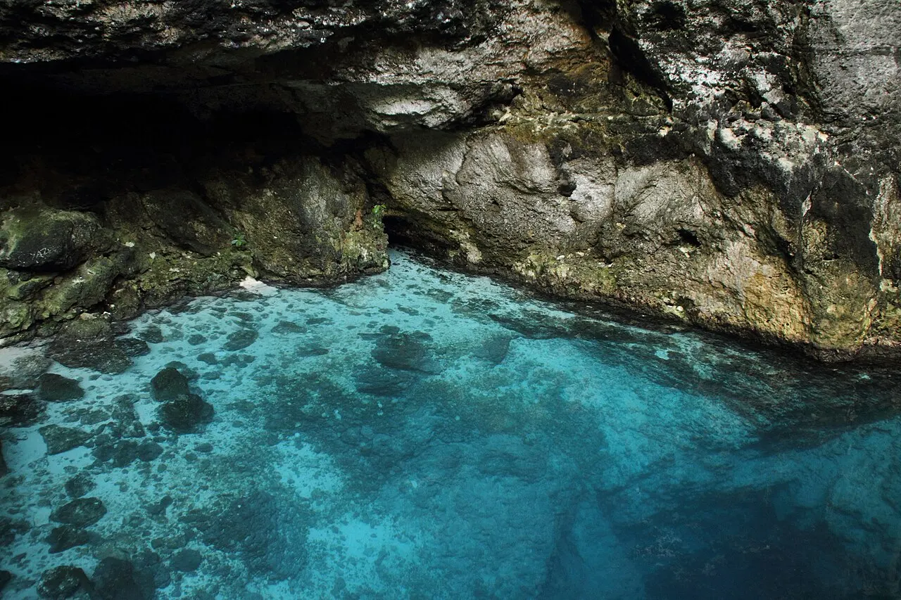 A água turquesa e transparente do Hoyo Azul, um cenote com o fundo de pedra visível, sob uma parede de rocha escura.