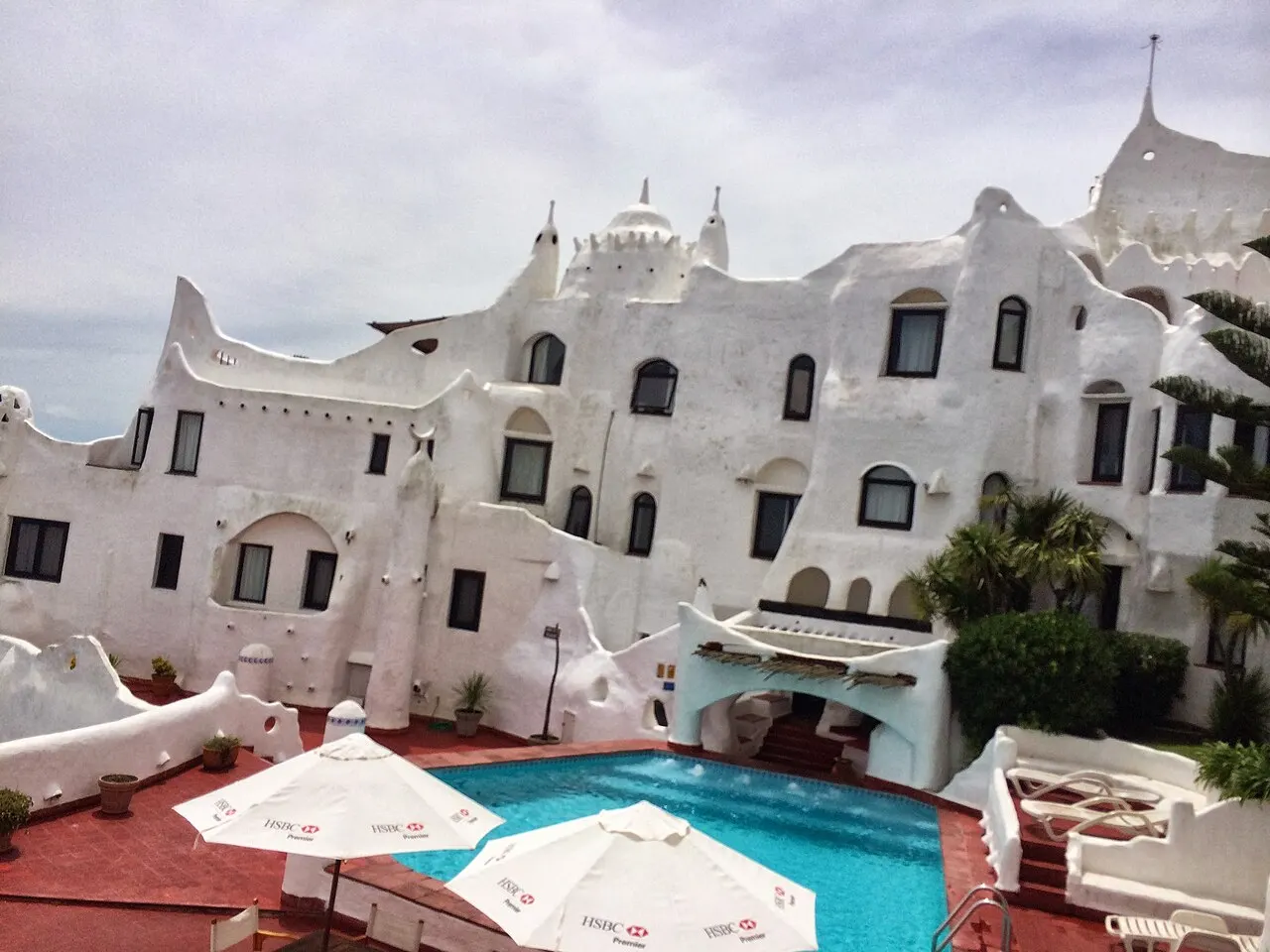 As paredes brancas e curvas de Casapueblo, com torres arredondadas e janelas irregulares, sobre uma piscina azul com guarda-sóis brancos.