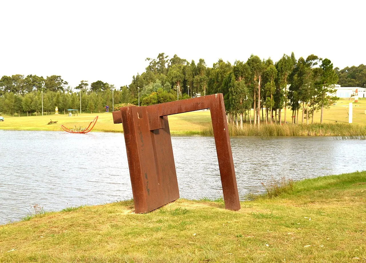 Uma escultura de aço enferrujado em forma de portal no gramado à beira do lago do parque da Fundación Pablo Atchugarry, com árvores ao fundo.