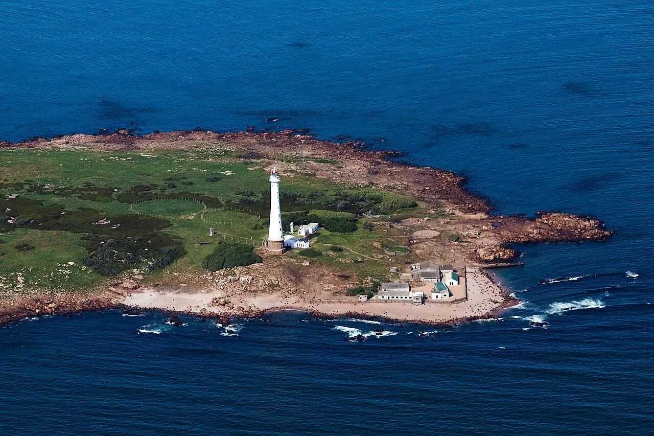 Vista aérea da Isla de Lobos, uma ilha baixa de pedra e capim no mar azul-escuro, com o farol branco e as construções do faroleiro.