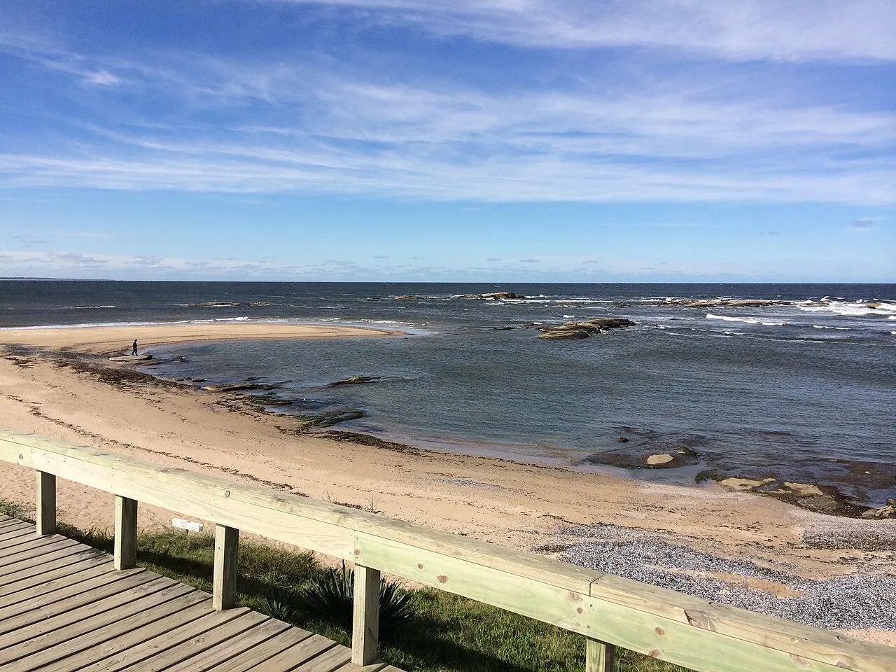 Uma passarela de madeira desce para a praia de José Ignacio, com areia clara, pedras na beira e o mar sob céu com nuvens finas.