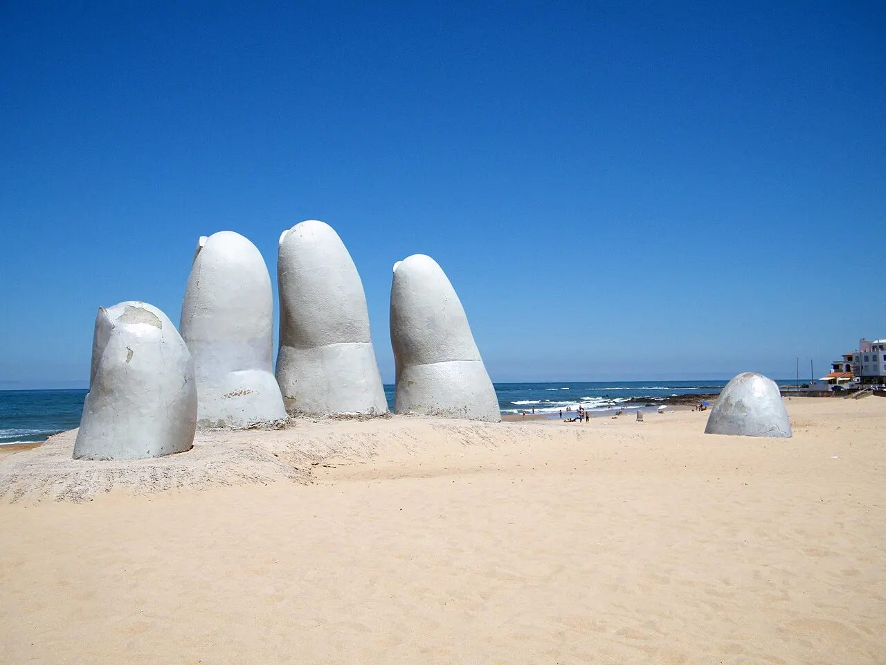 A escultura La Mano: cinco dedos de concreto cinza-claro saindo da areia da Playa Brava, com o mar e o céu azul atrás.