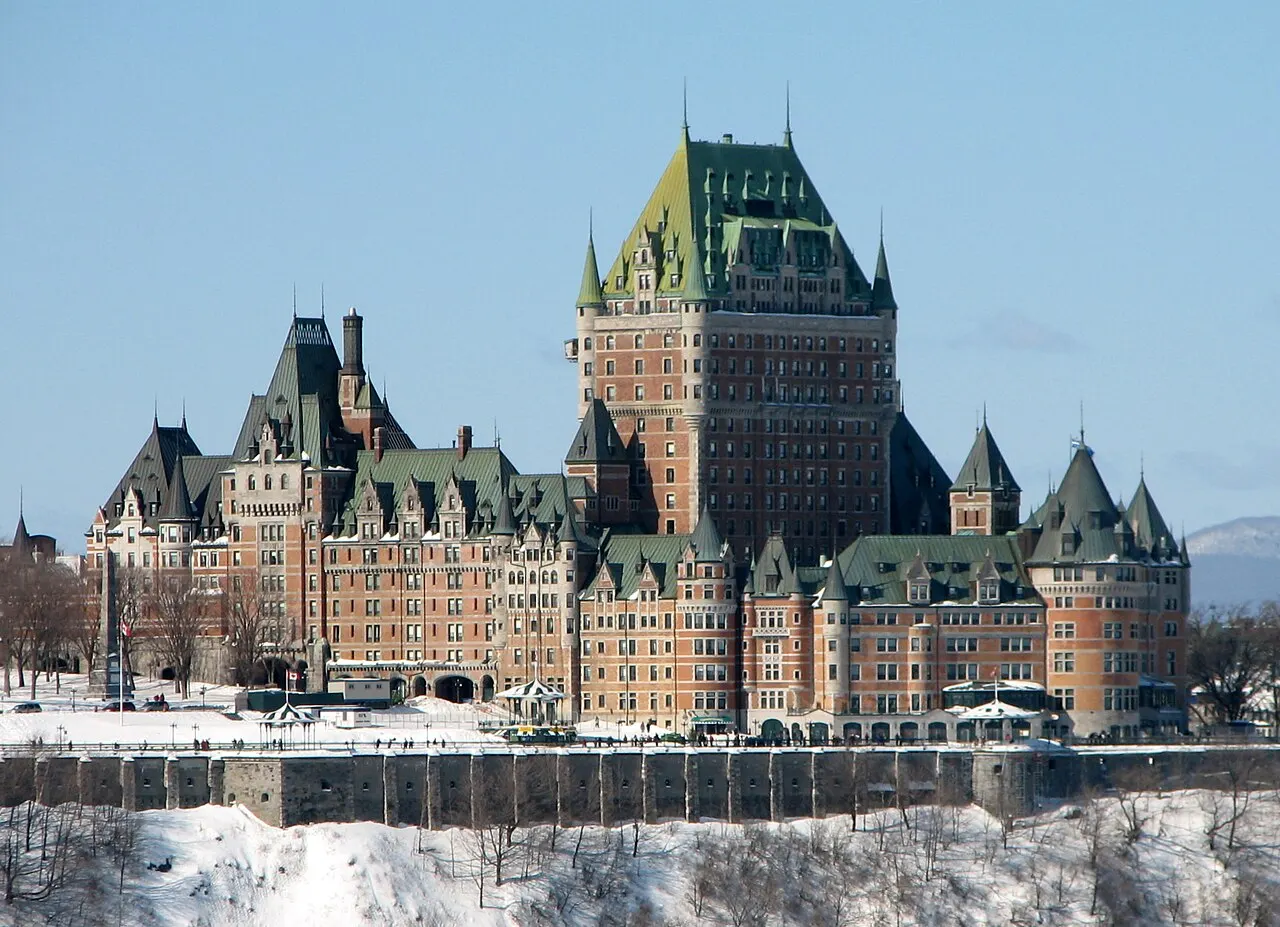O Château Frontenac, de tijolo e telhados verdes pontiagudos, com a torre central alta, visto de longe sobre a muralha e a encosta coberta de neve.