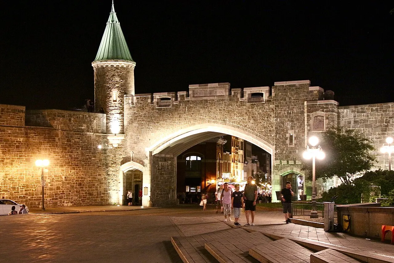 A Porte Saint-Jean à noite, um arco de pedra na muralha com ameias e uma torre de telhado verde em ponta, iluminada, com algumas pessoas passando pela praça.