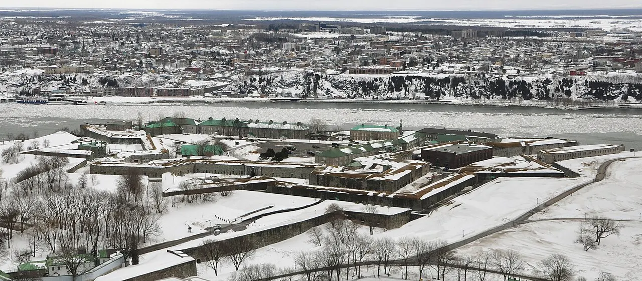 A Citadelle vista do alto no inverno, com os baluartes em forma de estrela cobertos de neve, os prédios de telhado verde no centro e o rio São Lourenço atrás.