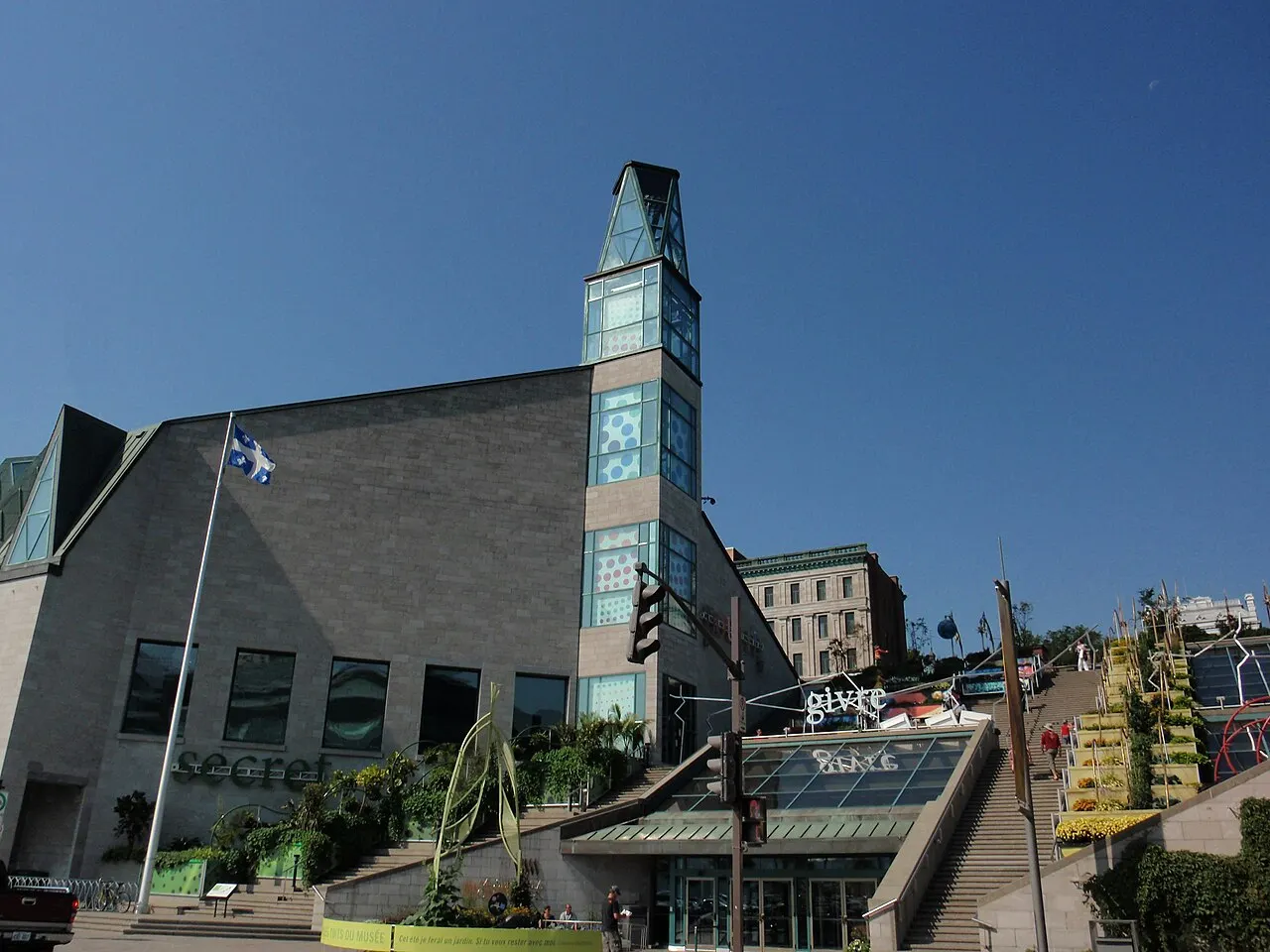 O prédio moderno do Musée de la civilisation, de pedra cinza com uma torre de vidro, a bandeira do Quebec no mastro e uma escadaria larga ao lado da entrada.