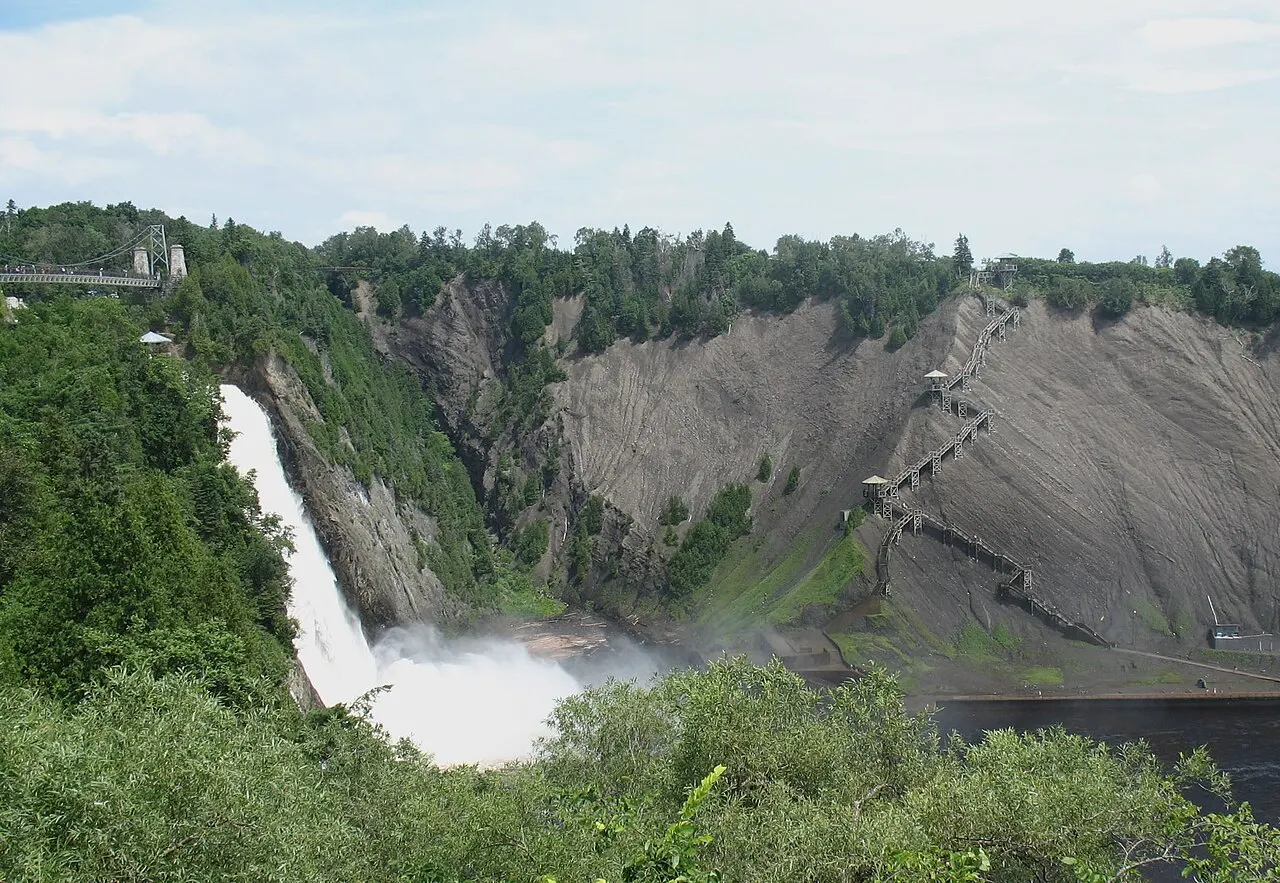 A queda-d'água da Montmorency despencando da borda da falésia numa nuvem de espuma, com a encosta de rocha cinza à direita, a escadaria de madeira subindo por ela e mata na frente.