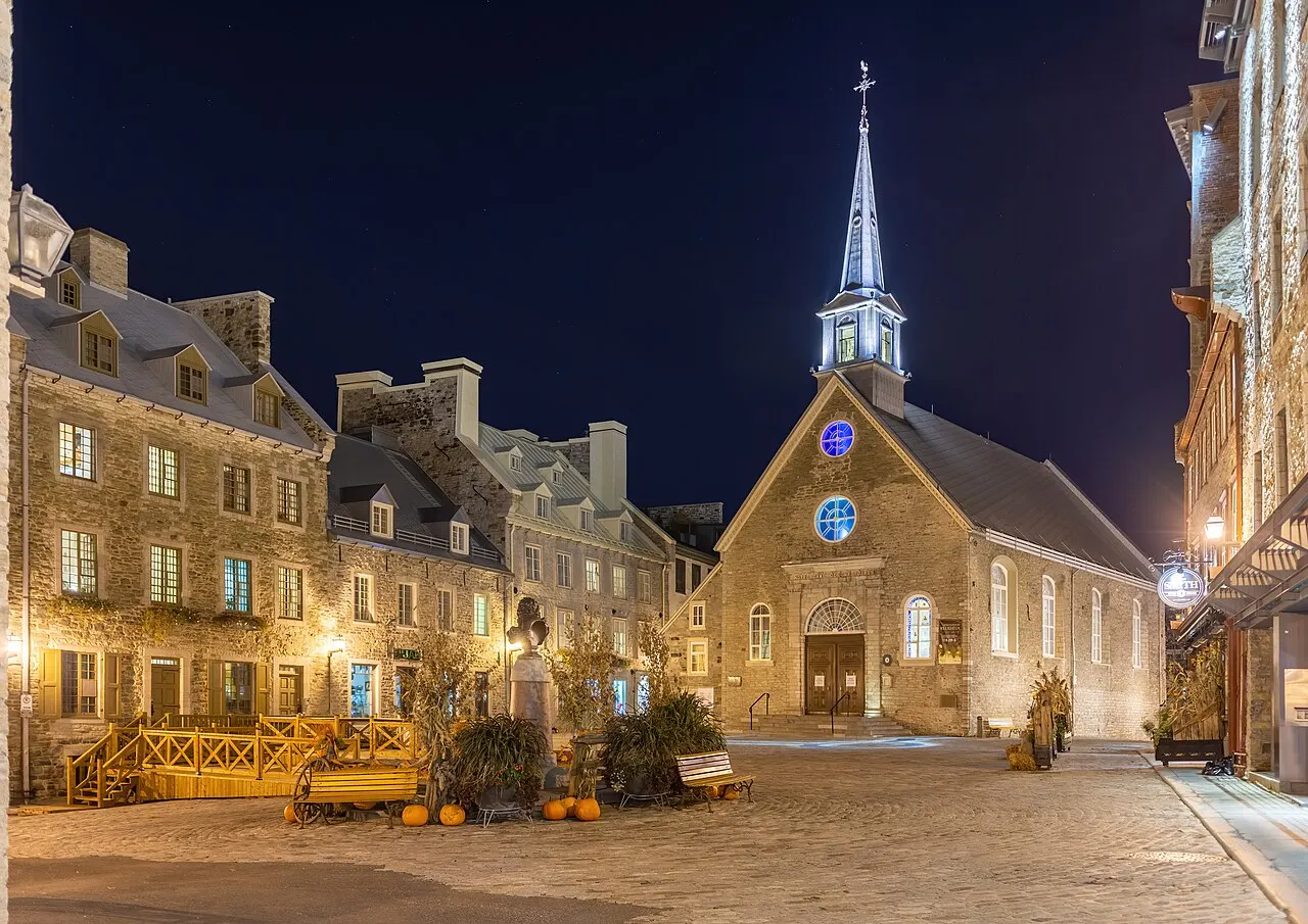 A Place-Royale à noite, com o calçamento de pedra, casas de pedra de telhado íngreme e, ao fundo, a pequena igreja Notre-Dame-des-Victoires com a torre iluminada.