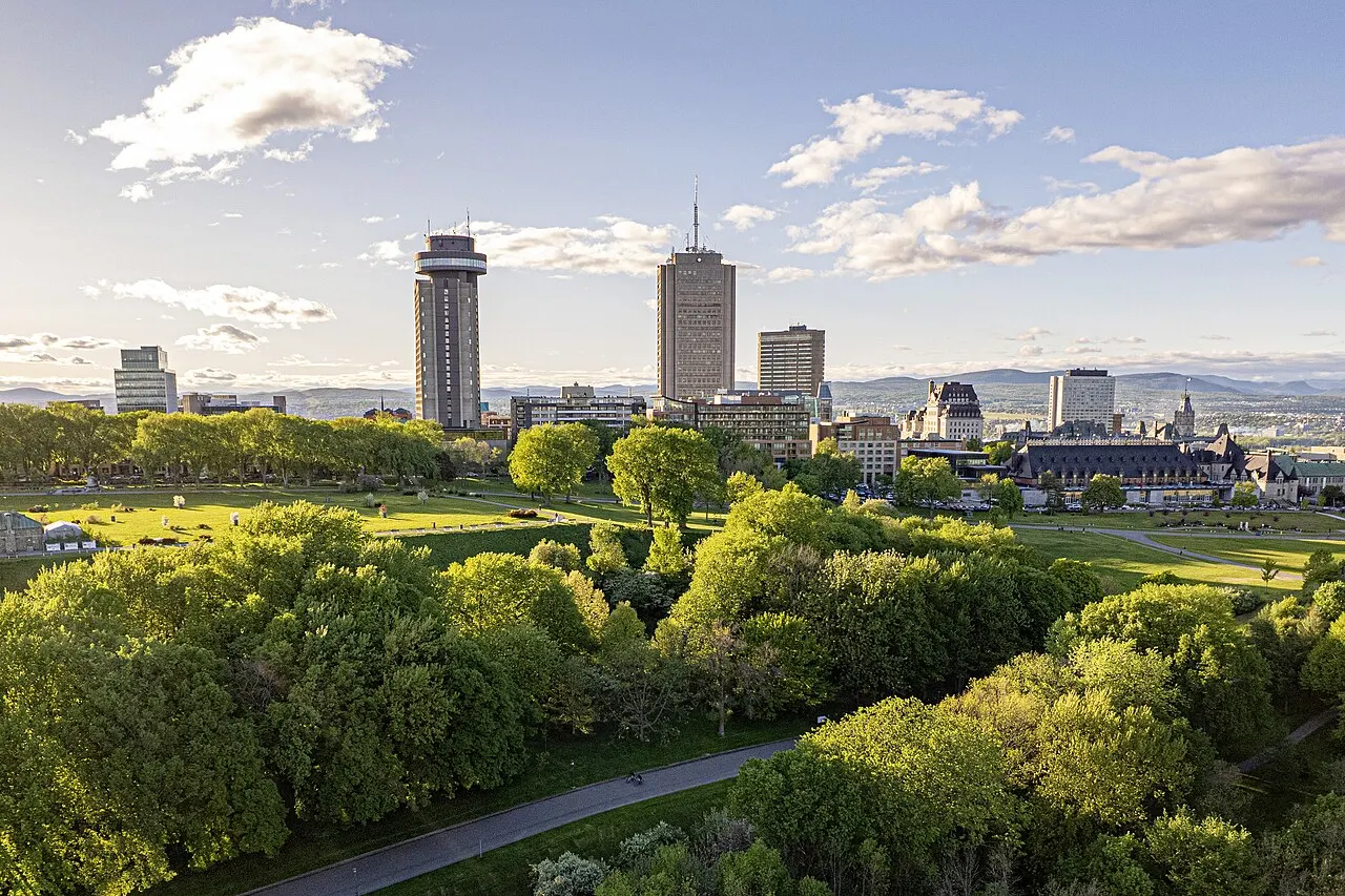 Vista aérea das Plaines d'Abraham, com árvores de copa verde em primeiro plano, gramados abertos e caminhos, e os prédios altos da cidade no horizonte.