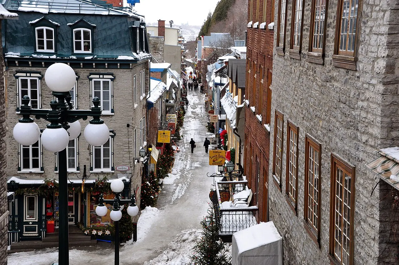A rua estreita do Petit-Champlain vista de cima, entre casas de pedra e tijolo, com neve na calçada, postes de globos brancos e pessoas caminhando ao longe.
