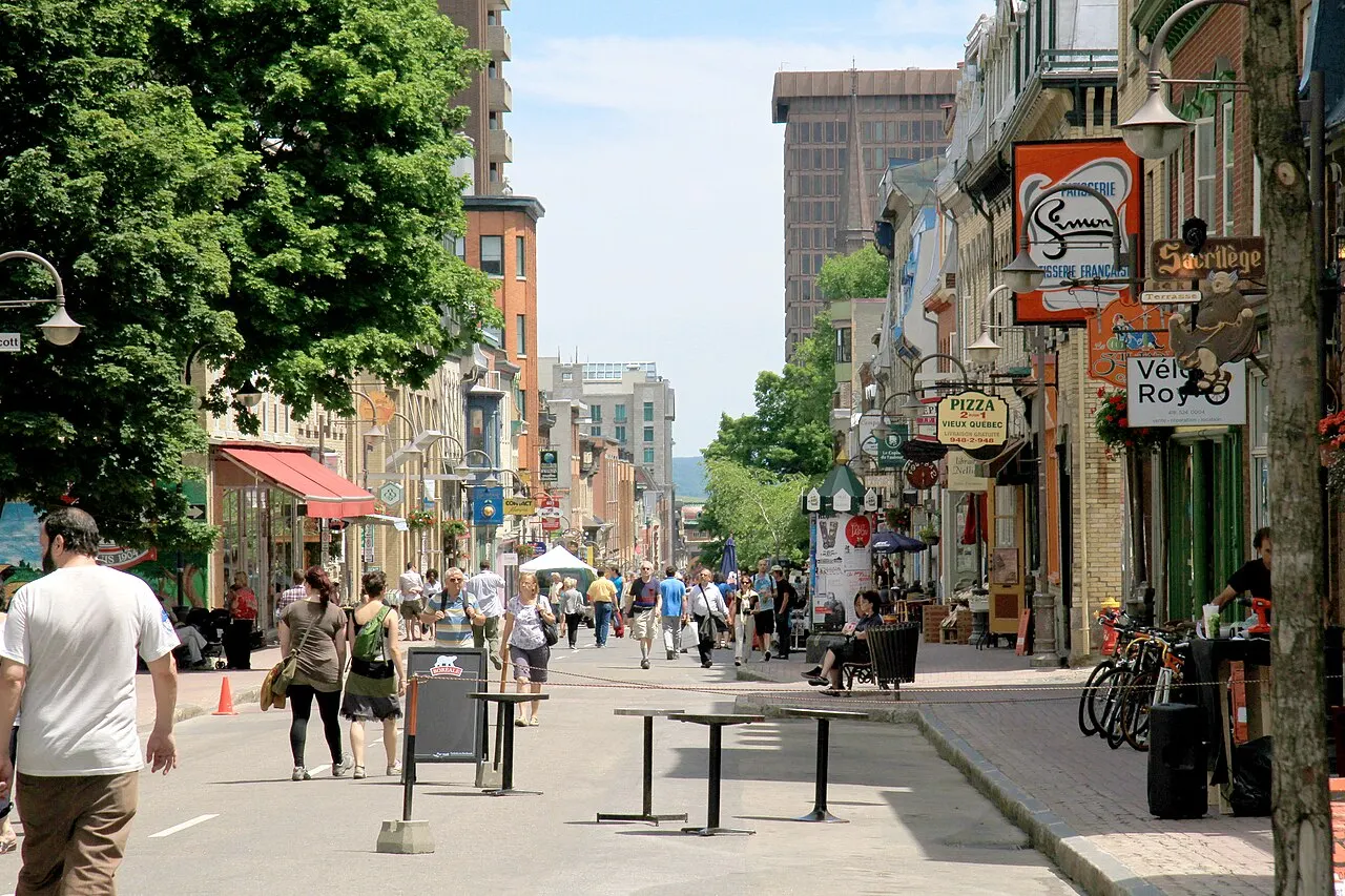A rue Saint-Jean num dia de sol, com pedestres andando no meio da rua, placas de lojas e restaurantes nas fachadas e árvores à esquerda.