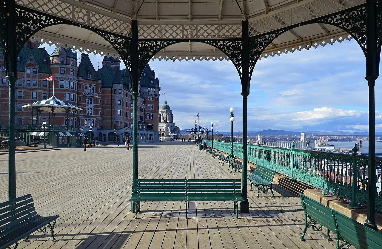 O calçadão de tábuas da Terrasse Dufferin visto de dentro de um quiosque de ferro, com bancos verdes, a grade sobre o rio à direita e o Château Frontenac ao fundo.